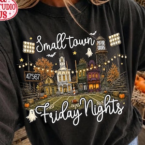 May include: A black t-shirt featuring a graphic print of a small town football field at night. The design includes a scoreboard reading 47567, a water tower labeled PETERSBURG, string lights, pumpkins, ghosts, and the text Small town Friday Nights.