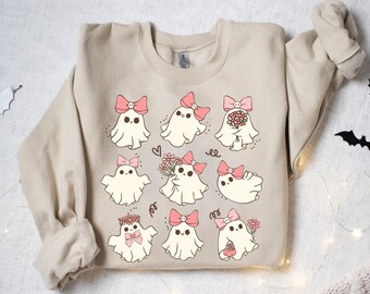 May include: A beige sweatshirt featuring a repeating pattern of cute cartoon ghosts with pink bows and floral accents. The ghosts have smiling faces and are in various poses. Black bat cutouts are scattered around the sweatshirt.
