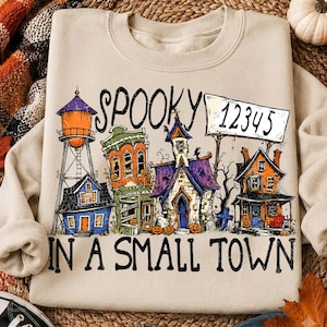 May include: A beige crewneck sweatshirt featuring a graphic print of a small town with a water tower, various buildings, and pumpkins. The text reads SPOOKY 12345 IN A SMALL TOWN in black lettering. The design includes orange, purple, and blue colors.