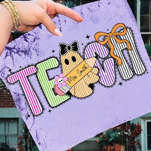 May include: A purple decorative square with the word "TEACH" in colorful letters. A ghost-shaped pencil with the name "Mrs. Smith" is featured. The design includes a black bow, an orange bow, and a flower.