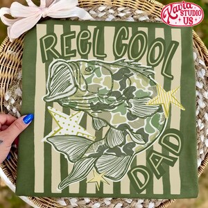 May include: Green t-shirt with the text "REEL COOL DAD" and a fish illustration in camouflage pattern. The design includes star accents. The shirt is displayed on a striped background.