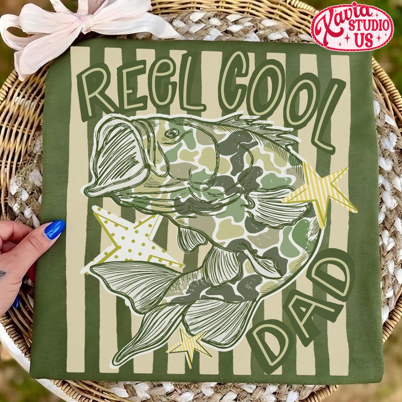 May include: Green t-shirt with the text "REEL COOL DAD" and a fish illustration in camouflage pattern. The design includes star accents. The shirt is displayed on a striped background.