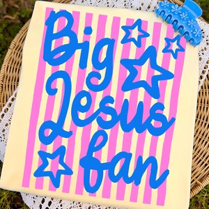 May include: A light yellow t-shirt with pink vertical stripes and the words "Big Jesus Fan" in blue script. Blue star graphics are scattered around the text. A blue floral hair clip is in the upper right corner.