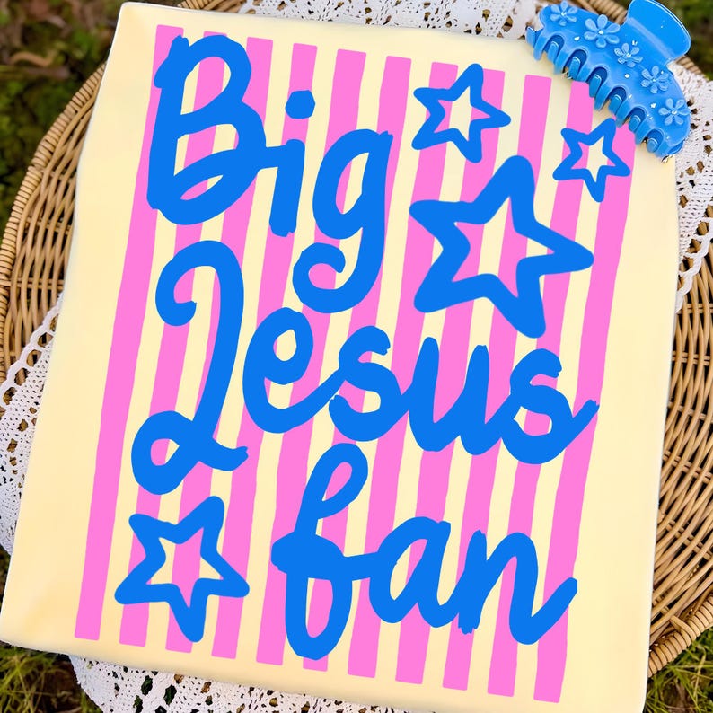 May include: A light yellow t-shirt with pink vertical stripes and the words "Big Jesus Fan" in blue script. Blue star graphics are scattered around the text. A blue floral hair clip is in the upper right corner.