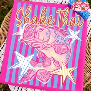 May include: A pink t-shirt with a graphic of a fish and the words "Shake That" in yellow script. The design includes blue and white stars and blue and pink stripes.
