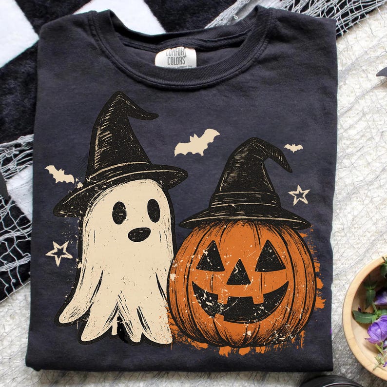 Op de afbeelding: Donkergrijs t-shirt met een Halloween-ontwerp. De afbeelding toont een geest met een heksenhoed en een pompoen, ook met een heksenhoed. Vleermuizen en sterren zijn verspreid rond het ontwerp. Het shirt heeft een ronde hals.