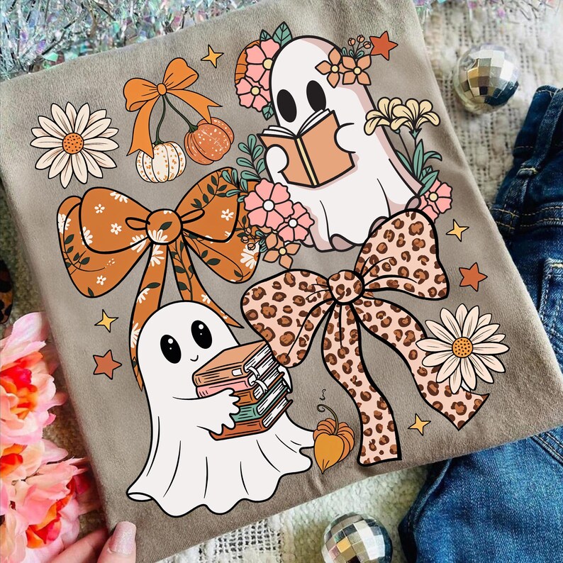 May include: A light gray t-shirt featuring a whimsical design with two cartoon ghosts, bows, flowers, and pumpkins. One ghost reads a book, while the other holds a stack of books. The bows have floral and leopard print patterns.