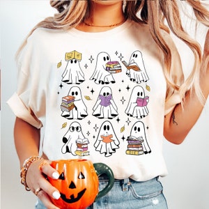 May include: Cream-colored t-shirt featuring a graphic of cartoon ghosts reading books. The ghosts are white with black eyes, and the books are various colors. The design includes yellow leaves and star accents, creating a fall or Halloween theme.