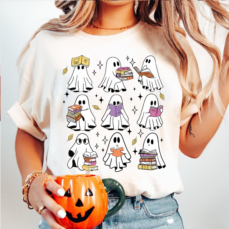May include: Cream-colored t-shirt featuring a graphic of cartoon ghosts reading books. The ghosts are white with black eyes, and the books are various colors. The design includes yellow leaves and star accents, creating a fall or Halloween theme.