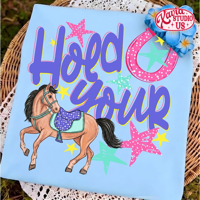 May include: Light blue t-shirt with a horse illustration and the words "Hold Your" in purple script. The design includes a pink horseshoe, stars, and a saddle on the horse. The shirt has a playful, equestrian theme.