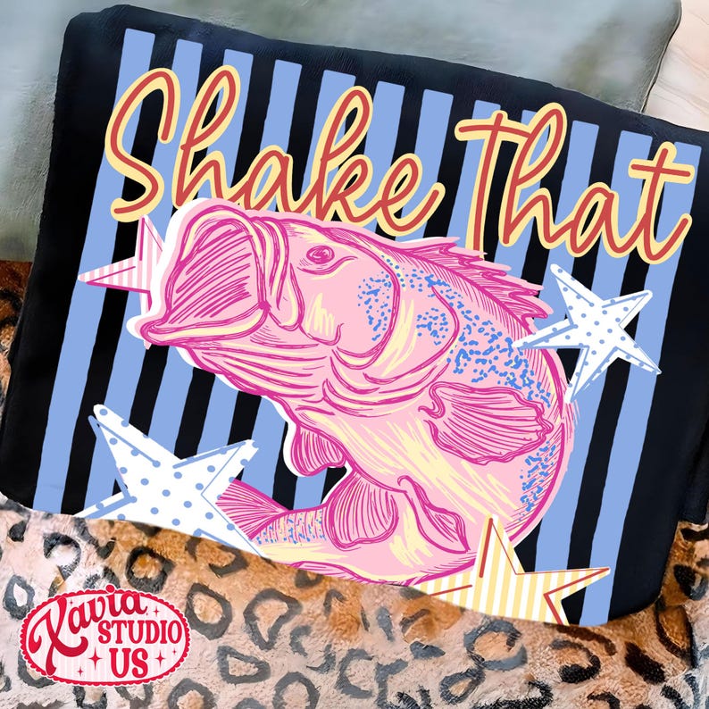 May include: A black throw pillow with a pink fish illustration and the words "Shake That" in a script font. The background has blue and black stripes with star accents. The pillow has a leopard print border.