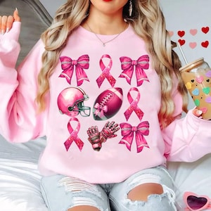May include: A light pink sweatshirt featuring a collection of pink football-themed graphics. The design includes pink bows, ribbons, a football helmet, a football, and football gloves. The graphics are arranged in a pattern across the front of the sweatshirt.