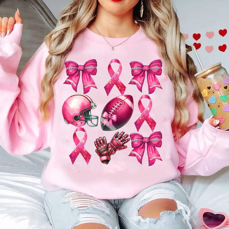 May include: A light pink sweatshirt featuring a collection of pink football-themed graphics. The design includes pink bows, ribbons, a football helmet, a football, and football gloves. The graphics are arranged in a pattern across the front of the sweatshirt.