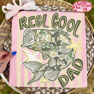 May include: Pink t-shirt with vertical stripes, featuring a graphic of a fish with camouflage pattern and the text "REEL COOL DAD" in green. The design includes star accents, perfect for a fishing enthusiast.