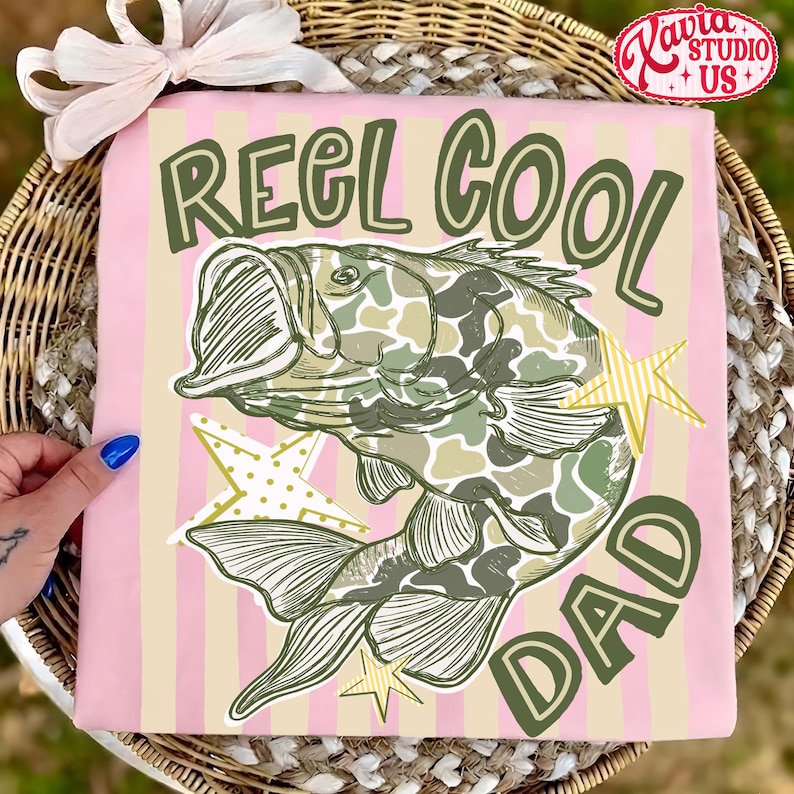 May include: Pink t-shirt with vertical stripes, featuring a graphic of a fish with camouflage pattern and the text "REEL COOL DAD" in green. The design includes star accents, perfect for a fishing enthusiast.