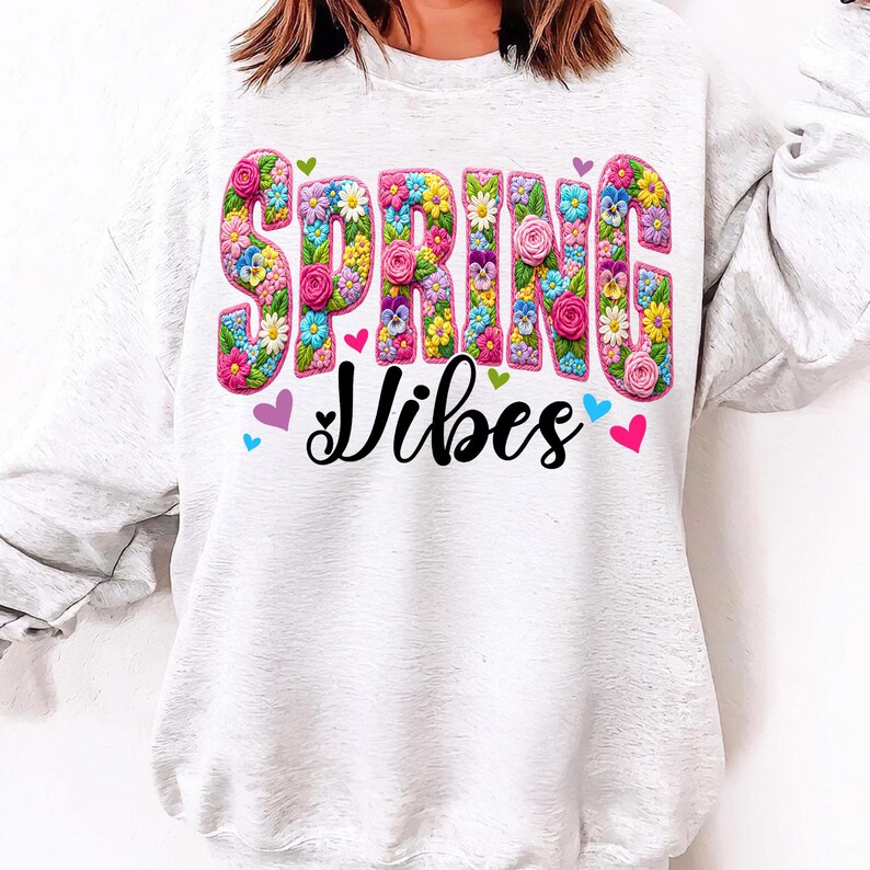 Puede incluir: Una sudadera gris claro con las palabras "SPRING VIBES" en letras grandes y coloridas llenas de flores. La palabra "Vibes" est&aacute; en una fuente cursiva. Peque&ntilde;os corazones de varios colores rodean el texto, creando un dise&ntilde;o alegre con tem&aacute;tica primaveral.