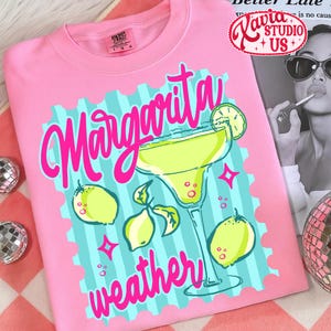 May include: Pink t-shirt with a margarita design. The design features the words "Margarita Weather" in pink script, a margarita glass, lemons, and stars. The shirt has a crew neck and short sleeves.