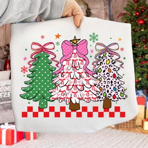 Puede incluir: Funda de cojín blanca con diseño navideño. El diseño presenta tres árboles de Navidad decorados: uno verde con lunares blancos, uno blanco con lazos rojos y otro con estampado de leopardo. Un borde a cuadros rojos y blancos en la parte inferior.