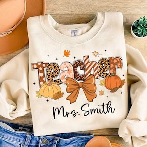 Può includere: Felpa color crema con la scritta "Teacher" in un design a tema autunnale, con zucche, foglie e un fiocco. Il nome "Mrs. Smith" è stampato sotto la parola "Teacher".