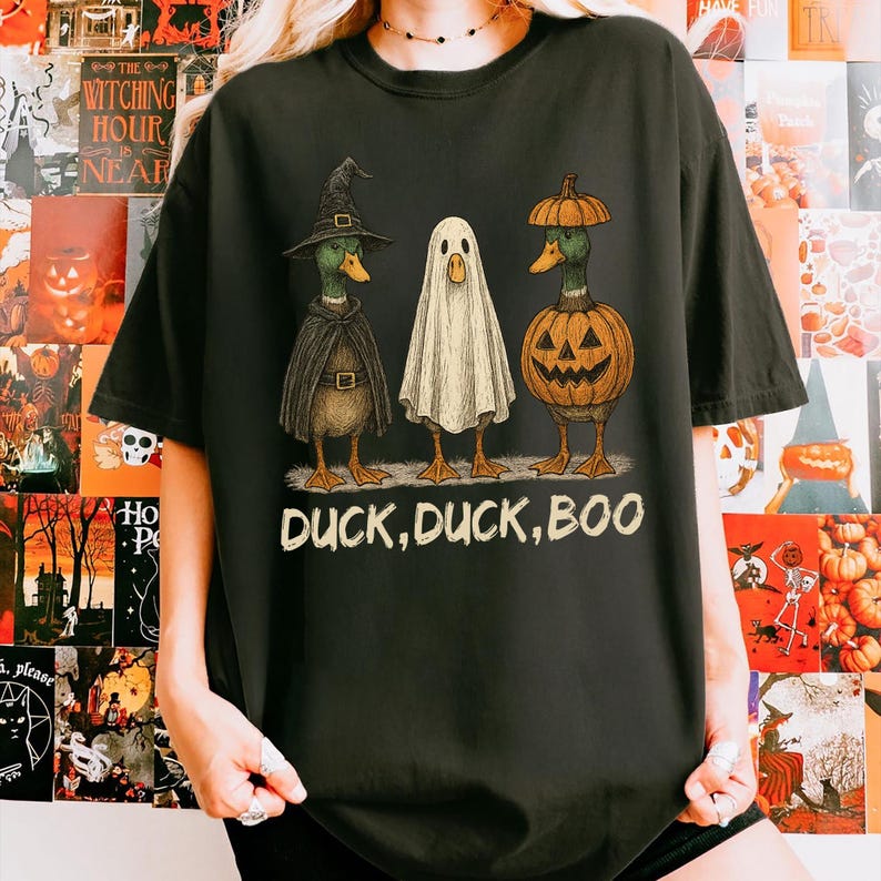 May include: Black t-shirt featuring a Halloween-themed graphic. The design includes three ducks dressed as a witch, a ghost, and a jack-o'-lantern. The text "DUCK, DUCK, BOO" is printed below the ducks. The shirt is a comfortable, casual style.