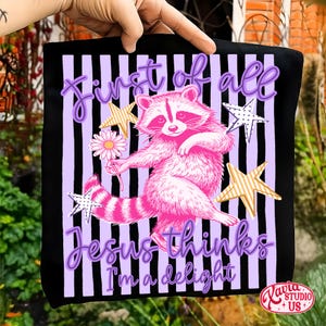 May include: Black tote bag with a pink raccoon illustration holding a daisy, surrounded by stars. The text reads "First of all Jesus thinks I'm a delight" in purple script. The background has vertical purple and black stripes.