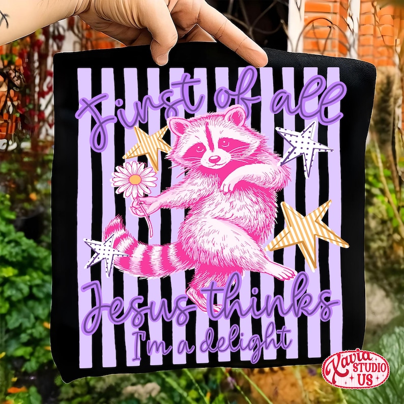 May include: Black tote bag with a pink raccoon illustration holding a daisy, surrounded by stars. The text reads "First of all Jesus thinks I'm a delight" in purple script. The background has vertical purple and black stripes.