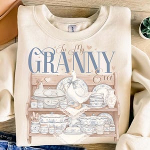 May include: Cream-colored sweatshirt with a graphic featuring a wooden shelf displaying blue and white china, a chicken, and a goose. The text "In My Granny Era" is written in blue script above the shelf, with small pink hearts.