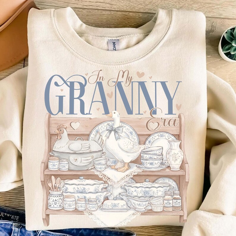 May include: Cream-colored sweatshirt with a graphic featuring a wooden shelf displaying blue and white china, a chicken, and a goose. The text "In My Granny Era" is written in blue script above the shelf, with small pink hearts.
