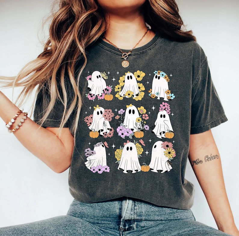 May include: Dark gray t-shirt featuring a grid of nine cartoon ghosts, each adorned with flowers and small pumpkins. The ghosts have black eyes and are surrounded by colorful floral arrangements. The shirt is a comfortable, casual style.