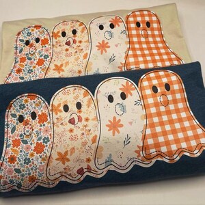 May include: Two folded t-shirts, one cream and one navy blue, each with a row of four ghost appliqués. The ghosts have black button eyes and various patterned designs, including floral and orange gingham, ideal for Halloween.