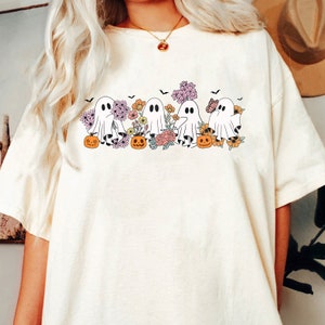 May include: A white t-shirt with a graphic of five ghosts holding pumpkins and flowers. The ghosts are white with black eyes and mouths. The pumpkins are orange with green stems. The flowers are pink, yellow, and orange.