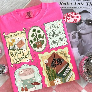 May include: A hot pink Comfort Colors t-shirt featuring a collage of book-themed illustrations. The design includes a coffee cup, potted plant, and the text "Fully Booked" and "One More Chapter". The shirt also has the logo "Kavia Studio US+".