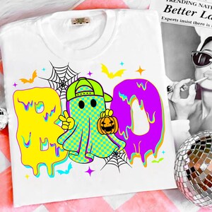 Puede incluir: Camiseta blanca con un gr&aacute;fico colorido de Halloween. El dise&ntilde;o presenta la palabra "BOO" en amarillo y morado, un fantasma a cuadros sosteniendo una calabaza, murci&eacute;lagos, estrellas y telara&ntilde;as.