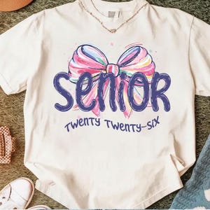 May include: Cream-colored t-shirt with a colorful bow graphic and the word "SENIOR" in dark blue. Below the word "SENIOR" is the text "TWENTY TWENTY-SIX" in a script font. The shirt has short sleeves.