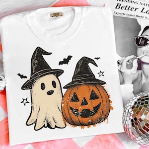Op de afbeelding: Wit T-shirt met een Halloween-ontwerp. De afbeelding toont een geest en een pompoen, beide met heksenhoeden. Het ontwerp bevat ook vleermuizen en sterren. Het T-shirt is gemaakt van een zacht materiaal.