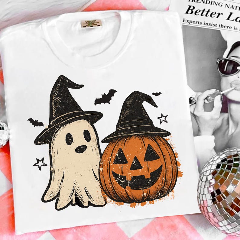 Op de afbeelding: Wit T-shirt met een Halloween-ontwerp. De afbeelding toont een geest en een pompoen, beide met heksenhoeden. Het ontwerp bevat ook vleermuizen en sterren. Het T-shirt is gemaakt van een zacht materiaal.