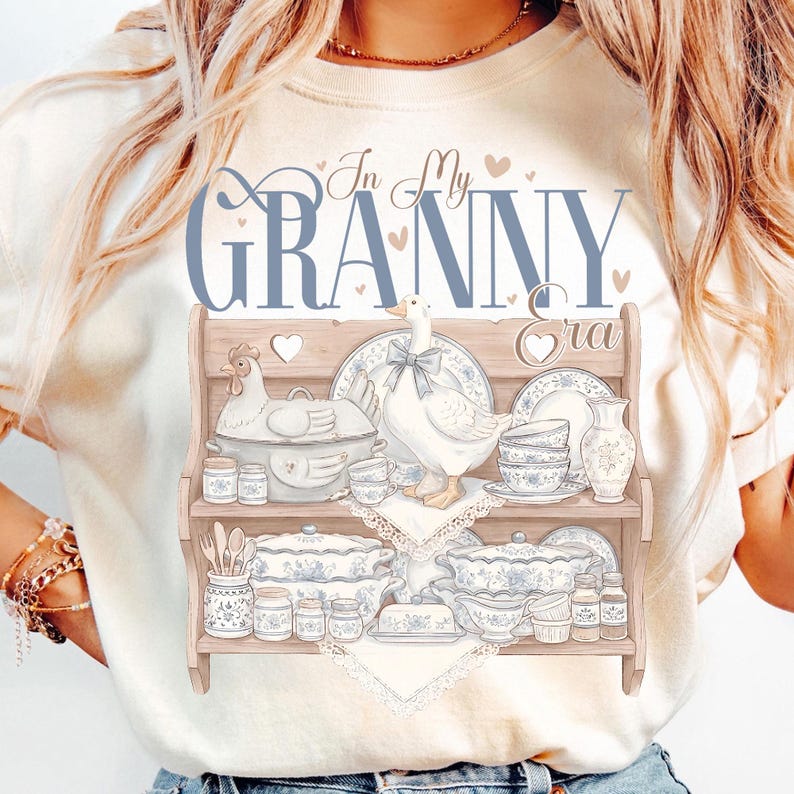 May include: Cream-colored t-shirt featuring a graphic of a wooden shelf displaying vintage-style dishes, a chicken, and a goose. The text "In My Granny Era" is written above the shelf in blue script. The design includes hearts and a lace doily.