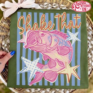May include: Olive green t-shirt with a pink bass illustration and the text "Shake That" in a playful font. The design includes blue and yellow stars, set against blue vertical stripes.