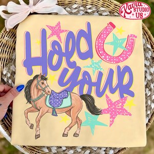 May include: A light yellow t-shirt with the words "Hold Your" in purple script, a pink horseshoe, and a cartoon horse with a saddle. Pink, blue, and yellow stars are scattered around the text and horse.