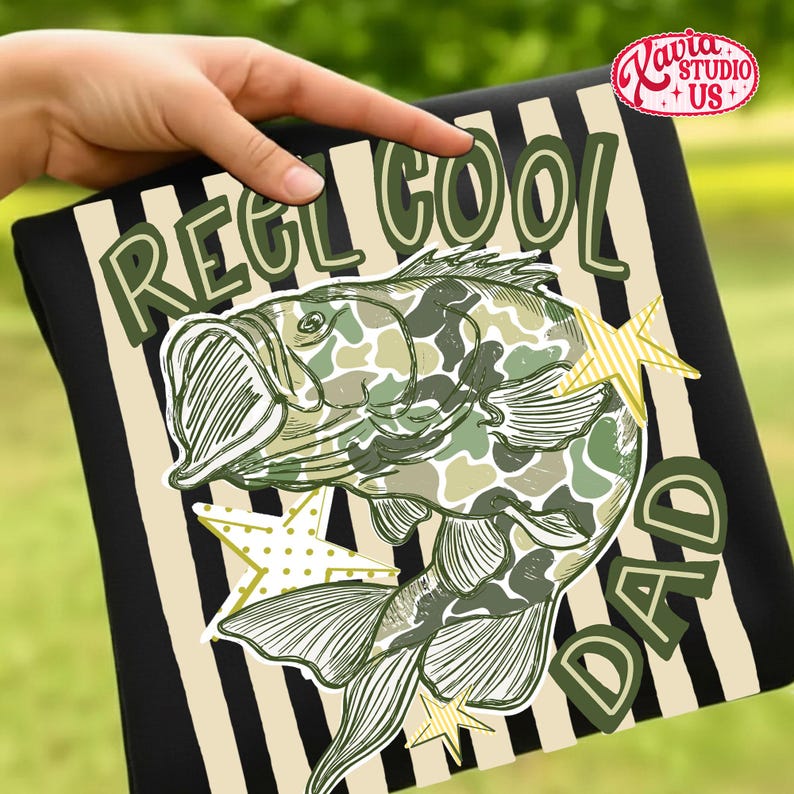 May include: Black t-shirt with a striped background design. The shirt features the text "REEL COOL DAD" with a fish illustration in camouflage pattern and star accents. The shirt is held by a hand.