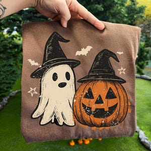 Könnte beinhalten: Eine braune Halloween-Tragetasche mit einem Geist und einem Kürbis mit Hexenhüten. Weiße Fledermäuse und Sterne sind um die Figuren herum verstreut. Der Geist ist cremefarben und der Kürbis ist orange.