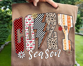 May include: A brown t-shirt with the words "COZY SEASON" in a colorful, decorative font. The letters feature patterns like cherries, leopard print, and a checkered design. The shirt is held up by a hand.