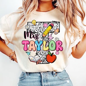 May include: A white t-shirt with a black and white polka dot design. The shirt features a large letter "T" with the text "Mrs. Taylor" in colorful bubble letters. The "T" is decorated with school-themed graphics, including a pencil, lightbulb, apple, book, and paint palette.
