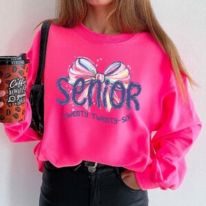 May include: A bright pink sweatshirt with the word "Senior" in blue script, adorned with a colorful bow graphic. The text "twenty twenty-six" is below the word "Senior". The sweatshirt is a vibrant, eye-catching piece.