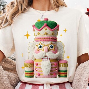 May include: Cream-colored t-shirt featuring a whimsical, detailed illustration of a nutcracker. The nutcracker is predominantly pink, green, and gold, with star-shaped glasses and a white beard. Gold stars are scattered around the design, creating a festive holiday theme.