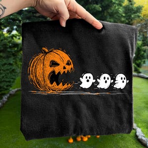 May include: Black Halloween-themed tote bag featuring an orange jack-o'-lantern with a carved face and three white cartoon ghosts. The design is a line drawing style, perfect for trick-or-treating or carrying groceries.