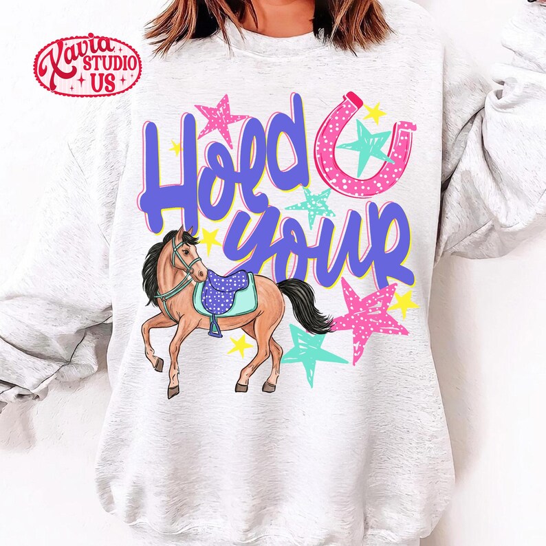 May include: A light gray sweatshirt with the words "Hold Your" in purple script, a pink horseshoe, and colorful stars. A cartoon horse with a blue saddle is also featured. The sweatshirt has a crew neck.