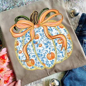 May include: A tan-colored t-shirt featuring a large, decorative pumpkin design. The pumpkin has a white background with blue floral patterns and small orange pumpkins. It is adorned with an orange bow and gold trim, perfect for fall.