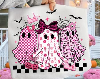 May include: A white Halloween-themed tote bag featuring three pink ghost characters with bows, patterns, and pink ribbon accents. The design includes bats, spiderwebs, and pink bra illustrations. A black and white checkered border completes the design.