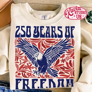 Puede incluir: Sudadera de color crema con un dise&ntilde;o patri&oacute;tico. La parte delantera presenta el texto "250 YEARS OF FREEDOM" en azul, con un &aacute;guila azul y detalles florales rojos. Un logotipo rojo y blanco est&aacute; en la esquina superior derecha.